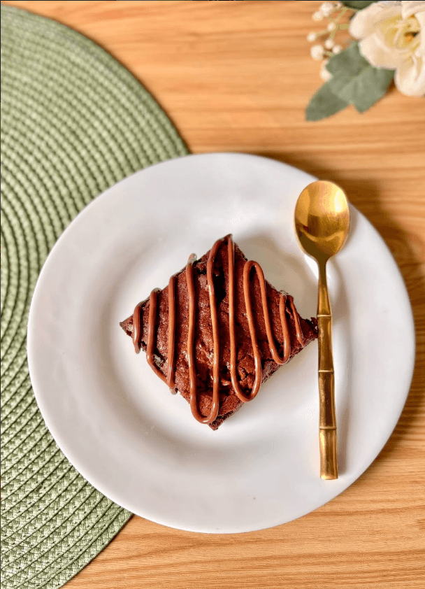 Fatia de brownie artesanal com calda de chocolate em prato branco e colher dourada da Krisllen Patrisserie